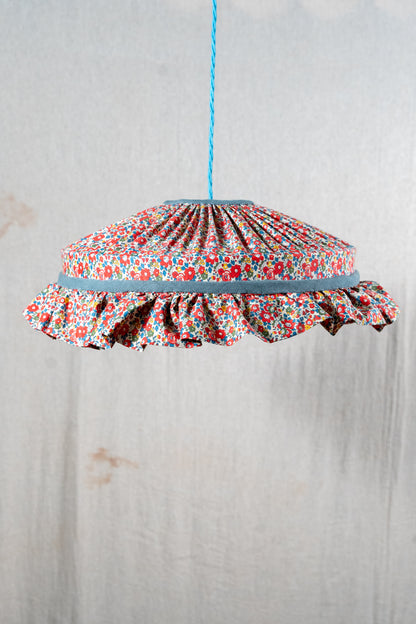 A hanging pendant light shade made of gathered floral fabric with a ruffled edge, suspended by a blue cord against a light textured wall.