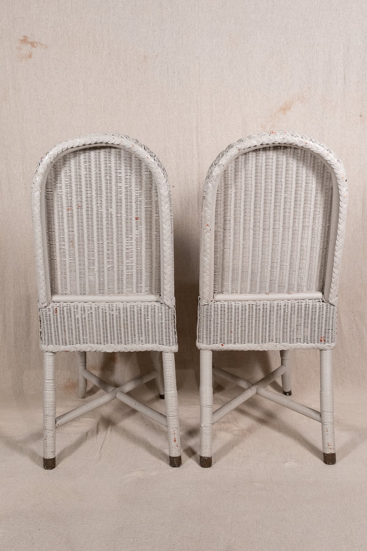 A pair of vintage white-washed wicker dining chairs with arched backs and crossed leg supports, set against a neutral beige backdrop.