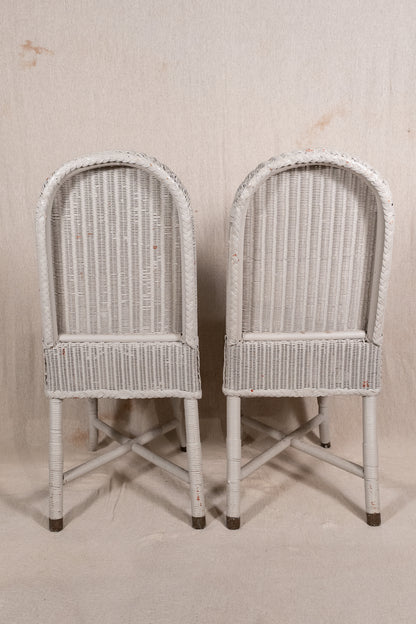 A pair of vintage white-washed wicker dining chairs with arched backs and crossed leg supports, set against a neutral beige backdrop.