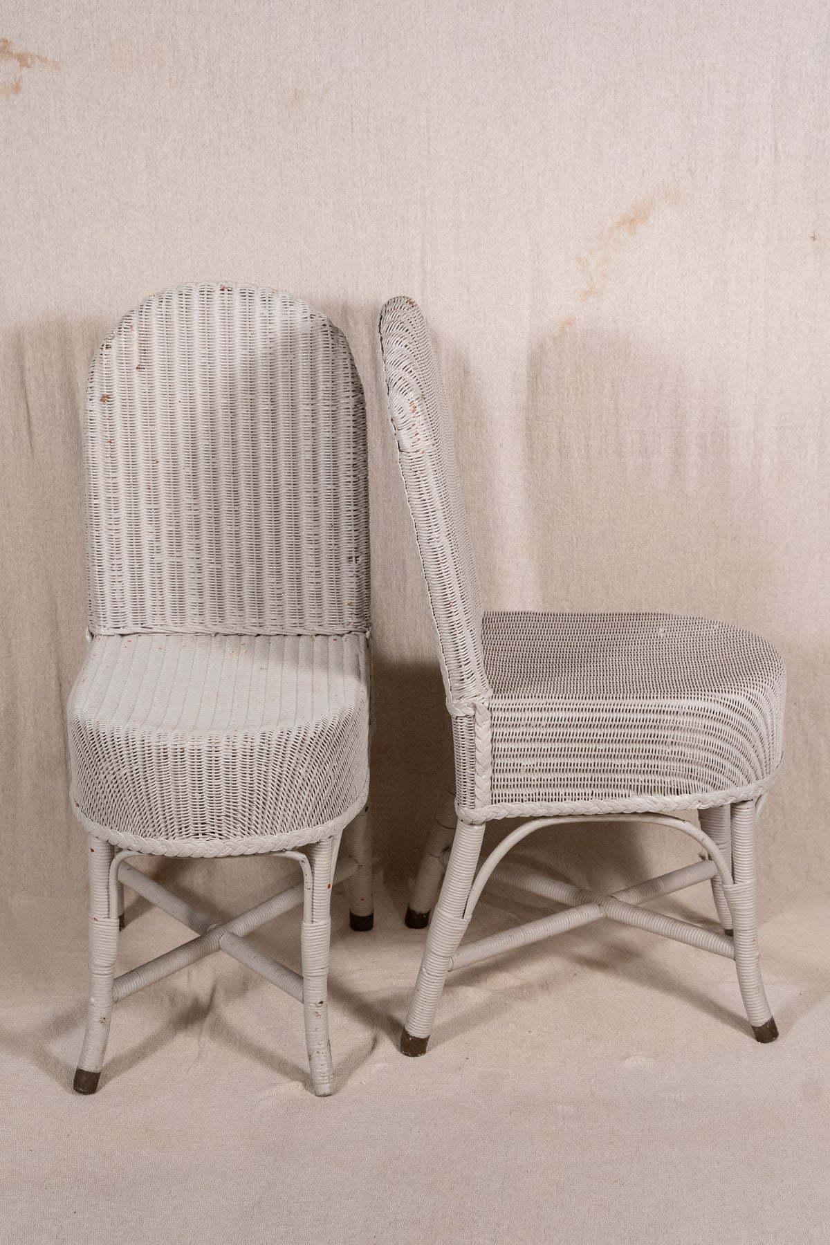 Two white wicker dining chairs with distressed finishes, shown against a neutral beige fabric backdrop.