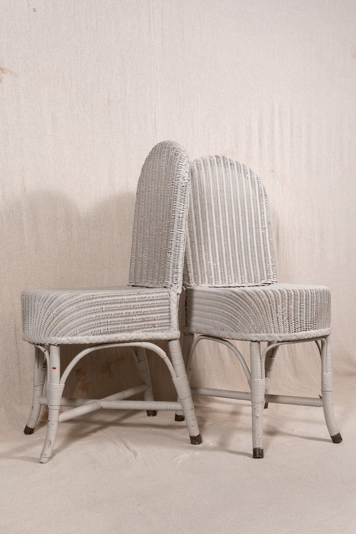 Two vintage white-washed wicker dining chairs with curved backs and striped backrests, set against a neutral textured beige backdrop.