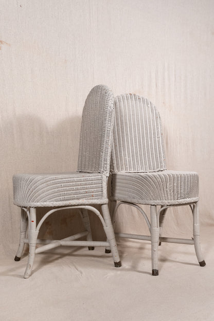 Two vintage white-washed wicker dining chairs with curved backs and striped backrests, set against a neutral textured beige backdrop.