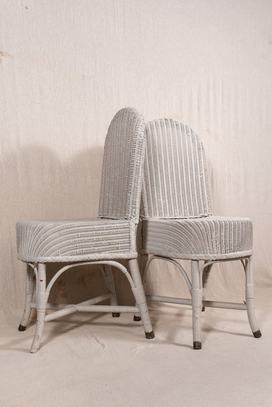 Two vintage white-washed wicker dining chairs with curved backs and striped backrests, set against a neutral textured beige backdrop.