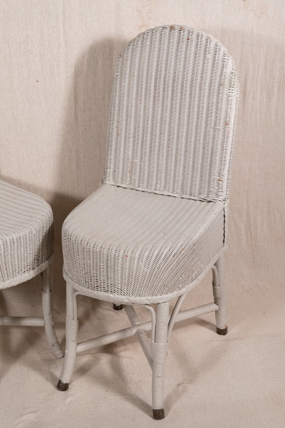 A white woven rattan dining chair with a high curved backrest and tapered legs, shown next to another matching chair against a neutral beige backdrop.