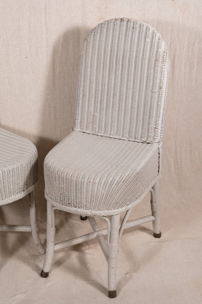 A white woven rattan dining chair with a high curved backrest and tapered legs, shown next to another matching chair against a neutral beige backdrop.