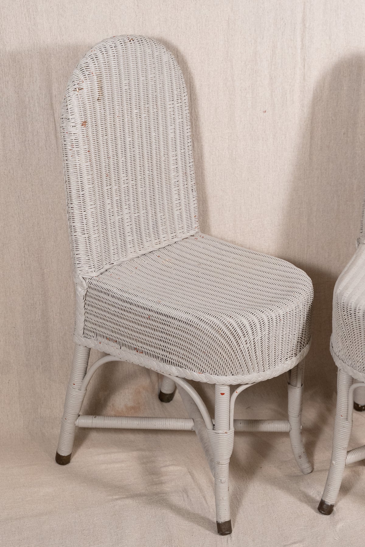 A white woven rattan or wicker dining chair with a curved backrest and tapered legs, shown against a light beige fabric backdrop. A second matching chair is partially visible on the right edge.