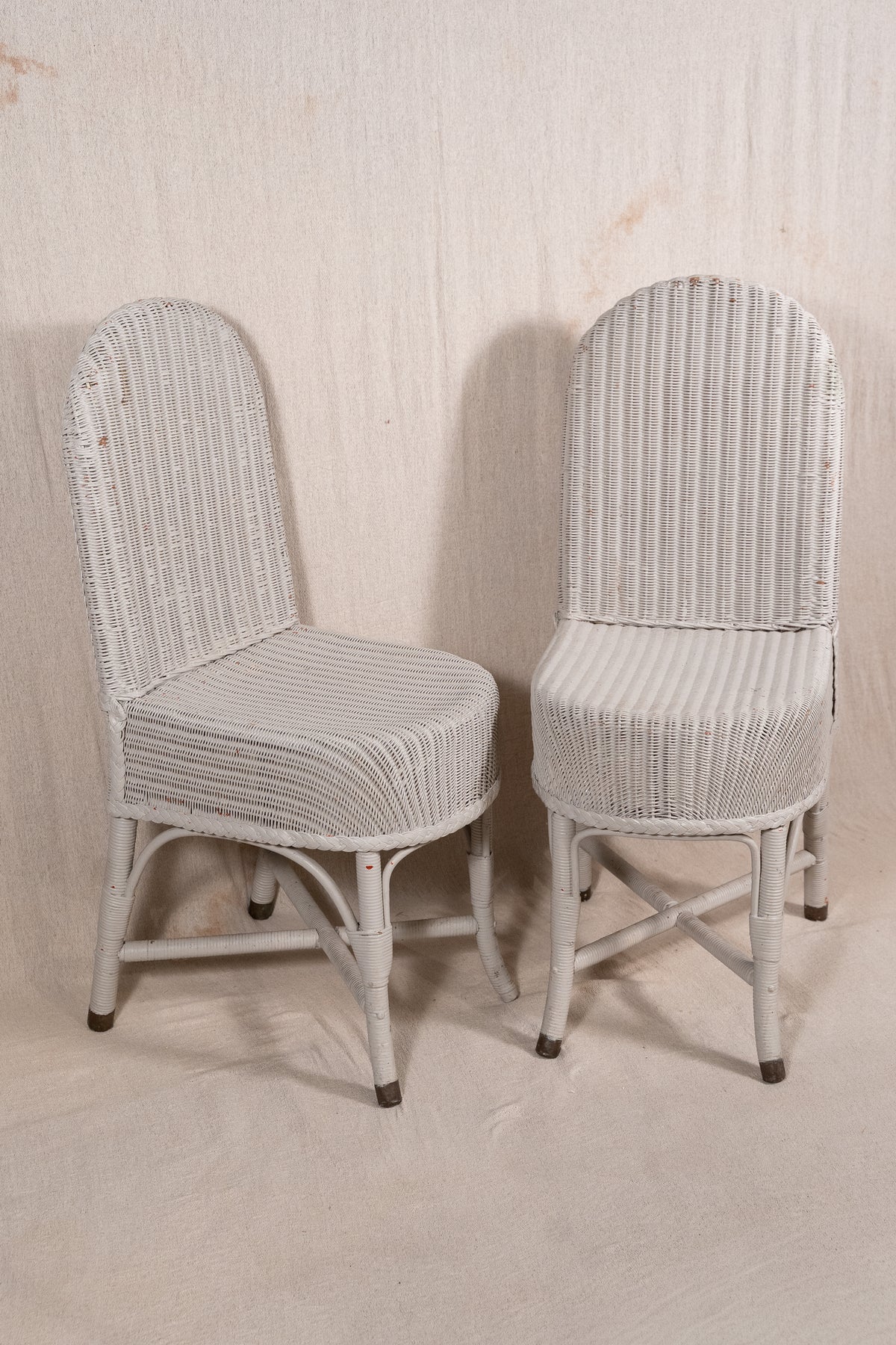 A pair of vintage white wicker dining chairs with curved backs and striped cushioned seats, set against a neutral textured beige backdrop.