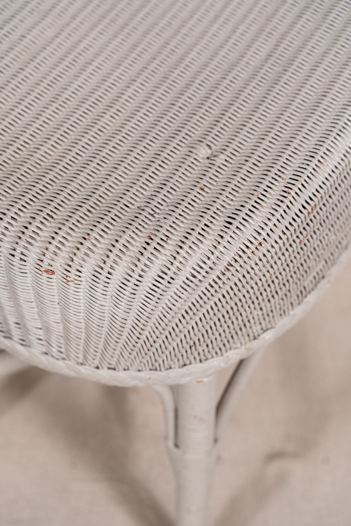 Close-up of a white woven wicker or rattan surface, likely part of a chair or stool, with a matching white frame leg visible below on a light neutral background.