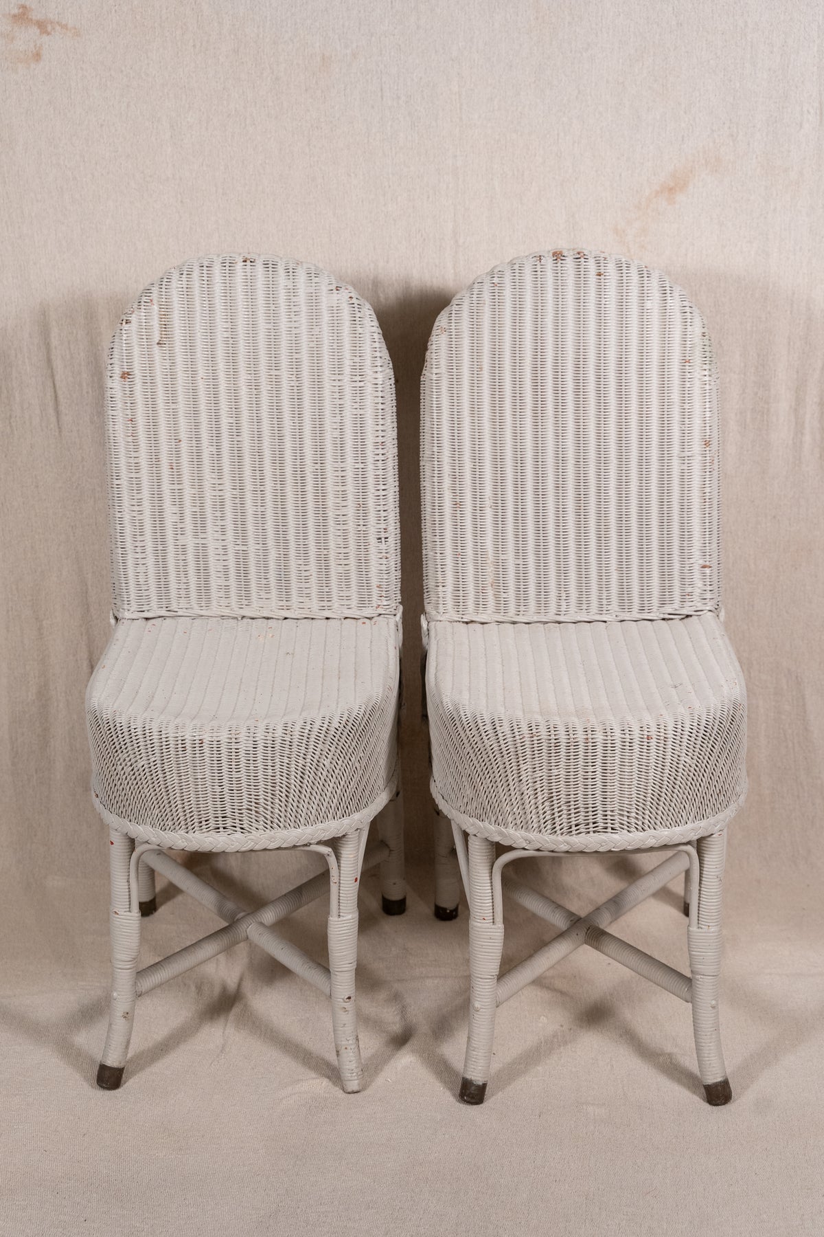 A pair of white wicker dining chairs with high arched backs and crossed leg frames, set against a neutral beige fabric backdrop.