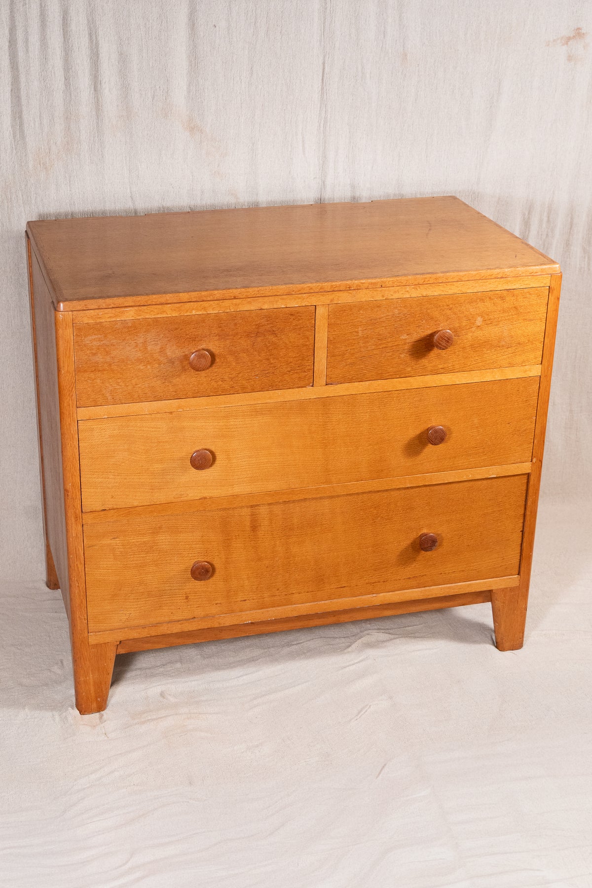 A mid-century modern wooden chest of drawers with a warm honey finish, featuring three top drawers with round knobs and two larger bottom drawers, photographed against a wrinkled white fabric backdrop.