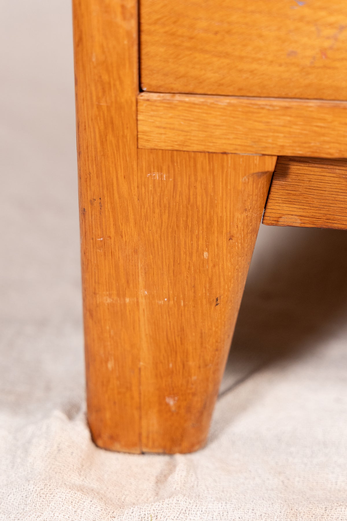 Close-up of a light brown wooden furniture leg, showing the wood grain and a portion of the drawer front, set against a neutral beige background.