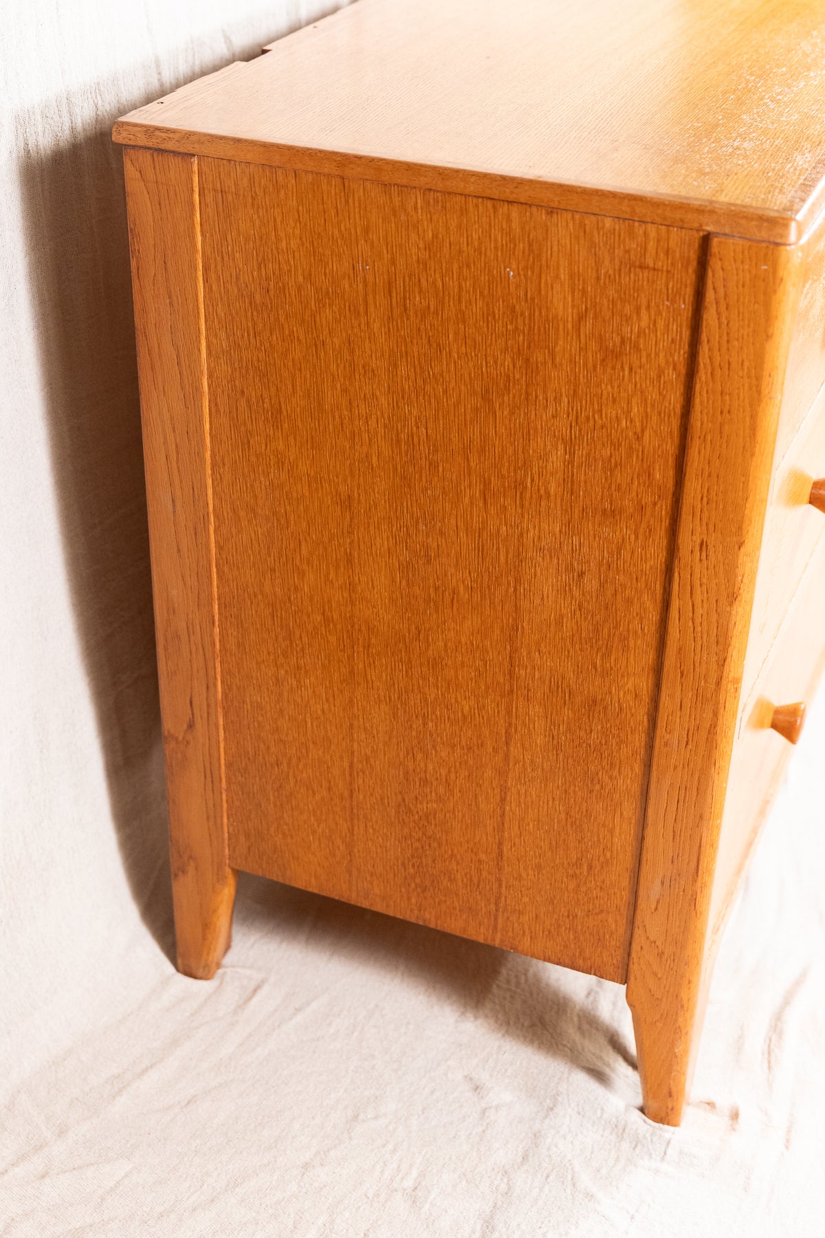 A close-up of a mid-century modern wooden cabinet or dresser in a warm honey oak finish, showing the wood grain and a small wooden drawer pull on the right side.