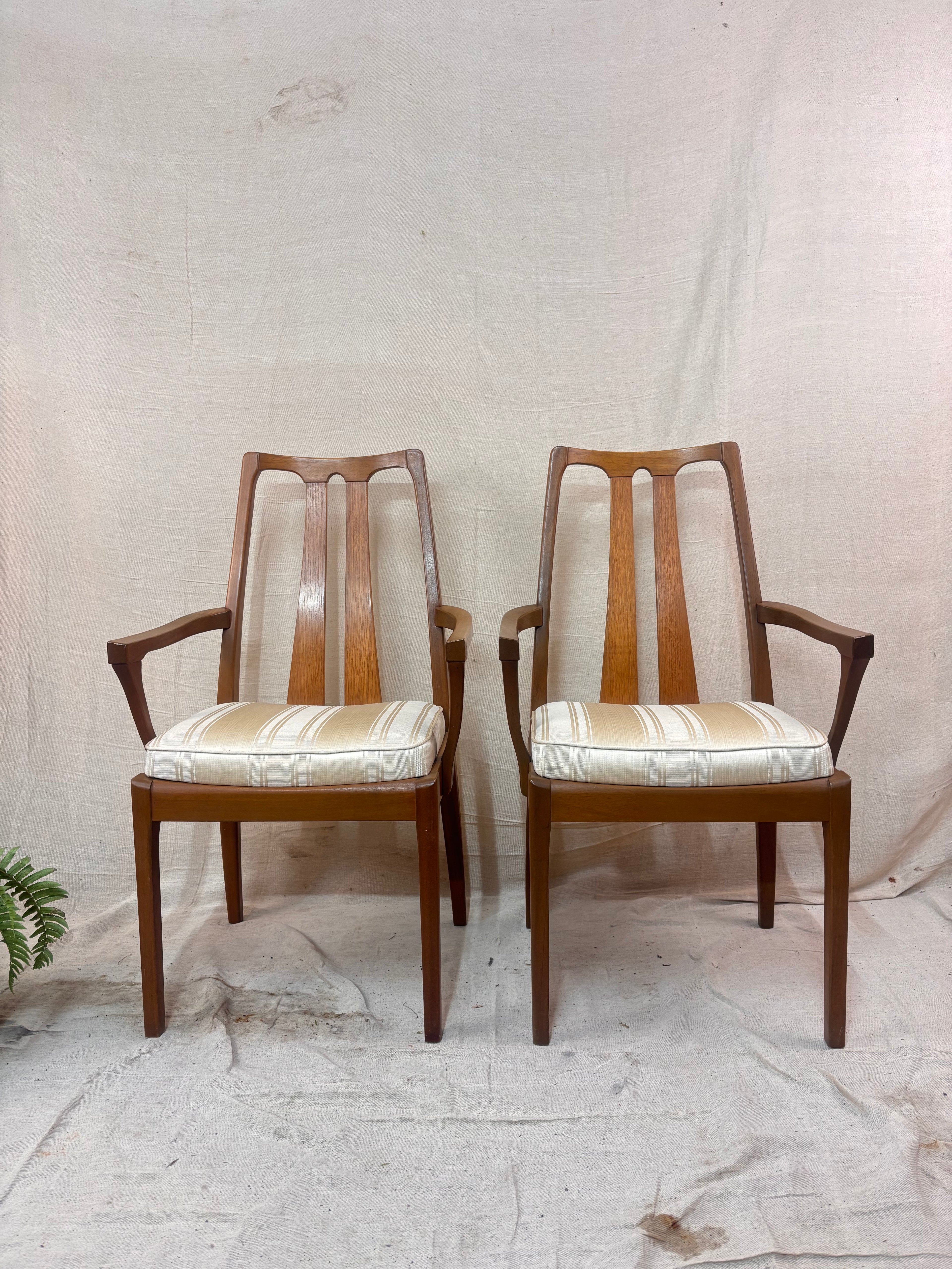 a pair of wooden dining chairs with arms and upholstered seats on a cream background