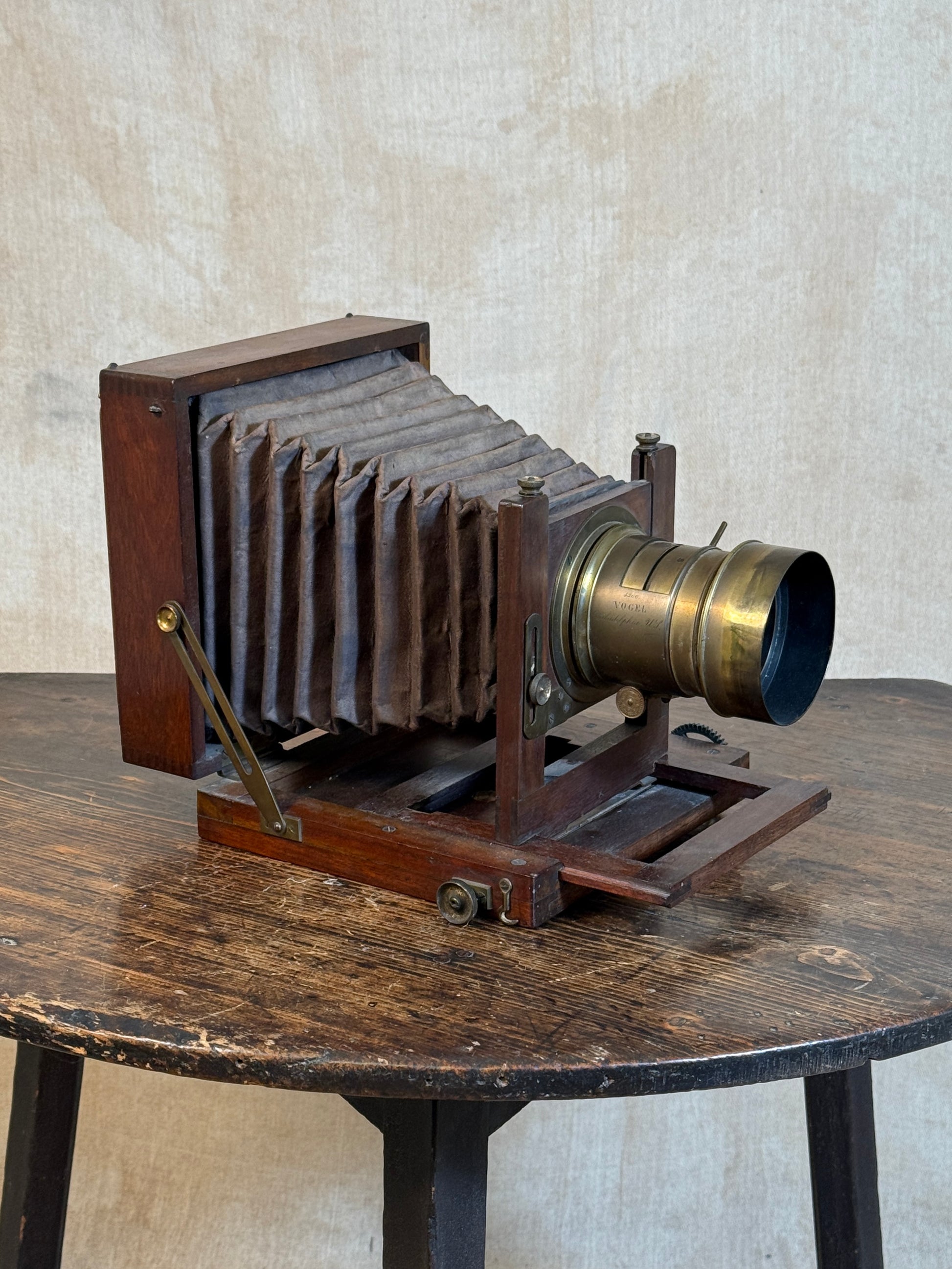 An antique folding camera with a dark wood body and bellows, a brass lens, and brass support arms, displayed on a rustic wooden table against a textured beige wall.