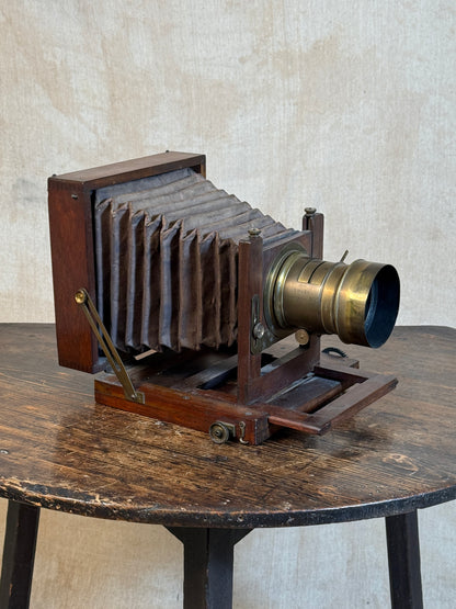 An antique folding camera with a dark wood body and bellows, a brass lens, and brass support arms, displayed on a rustic wooden table against a textured beige wall.