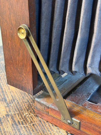 Close-up of a vintage folding camera with a wooden body, dark gray fabric bell, and brass hinge mechanism on a wooden surface.