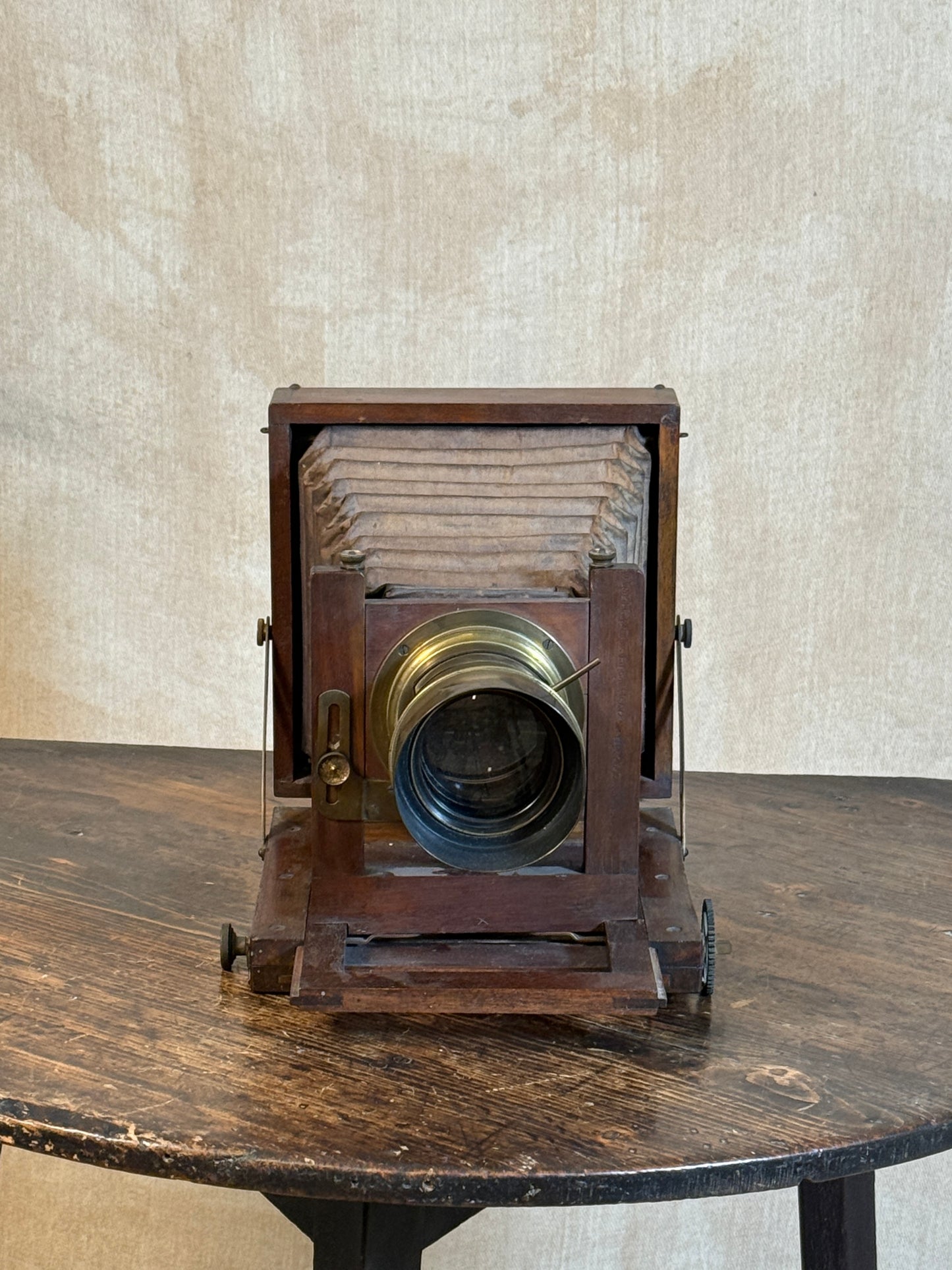 An antique wooden folding camera with a brass lens, displayed on a dark wooden table against a textured beige wall.