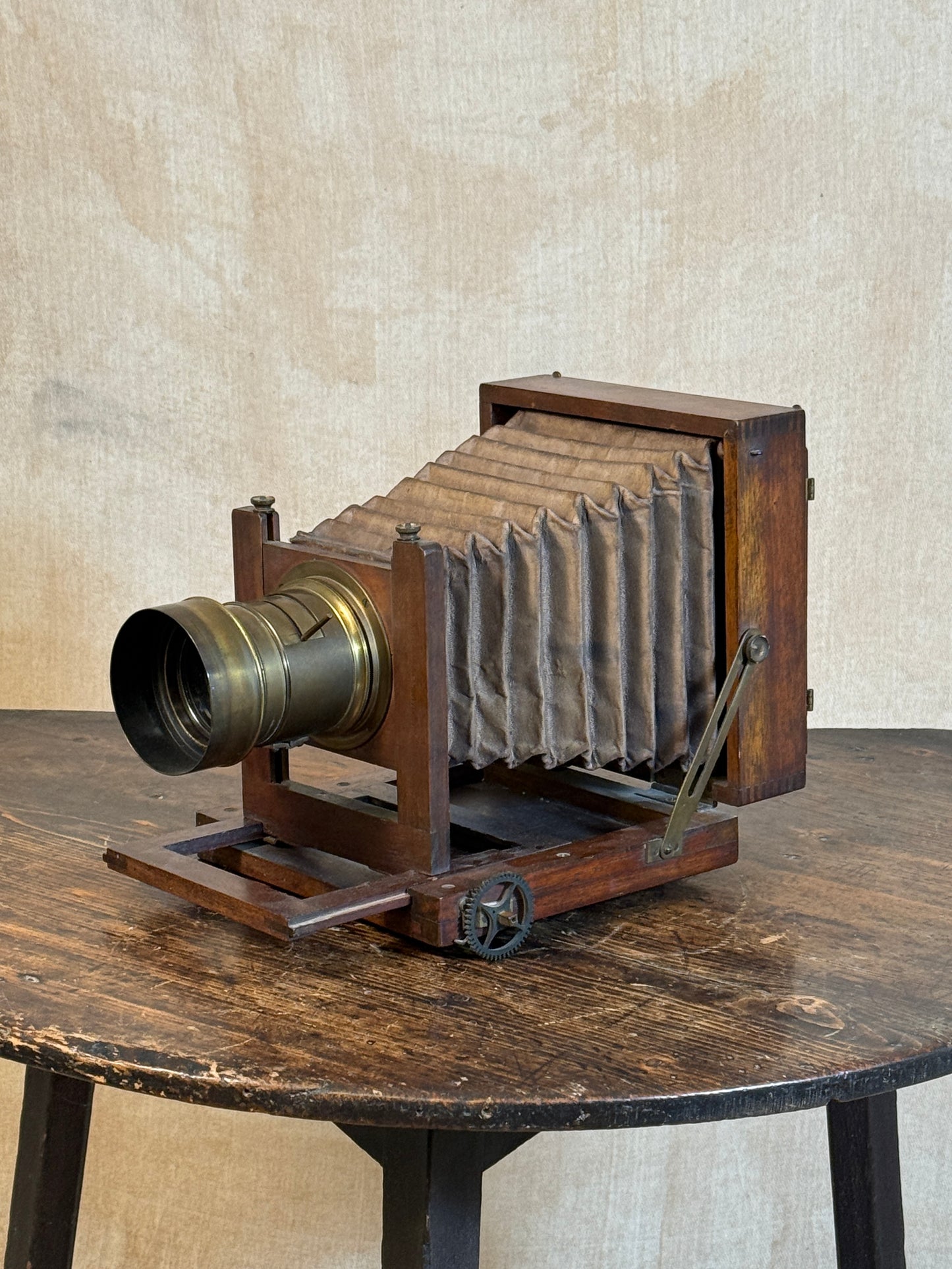 An antique wooden bellows camera with a brass lens, displayed on a rustic wooden table against a textured beige wall.