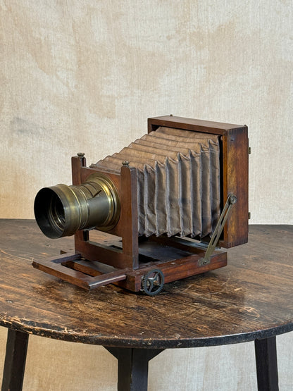 An antique wooden bellows camera with a brass lens, displayed on a rustic wooden table against a textured beige wall.