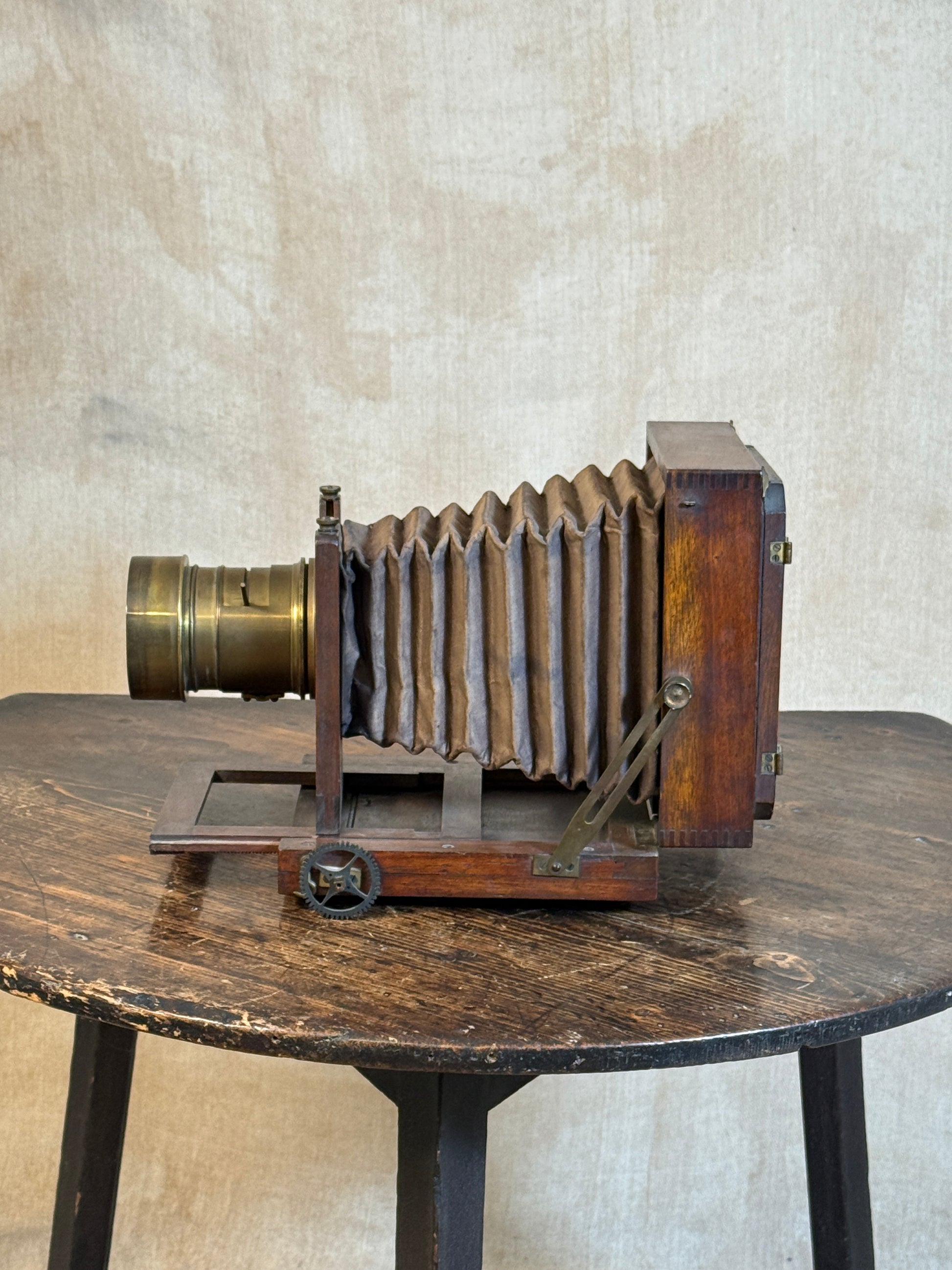 An antique folding camera with a dark wood body and bellows, displayed on a rustic wooden table against a textured beige wall.