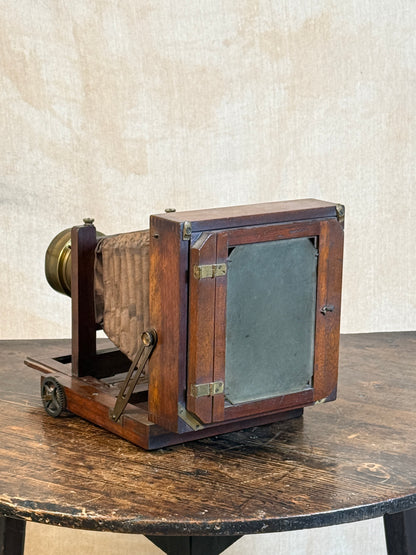 An antique wooden folding camera with brass hardware, displayed on a dark wooden table against a textured beige wall.