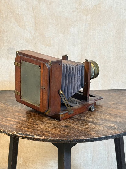 An antique wooden folding camera with a dark gray bellows and brass lens, displayed on a rustic wooden table against a textured beige backdrop.