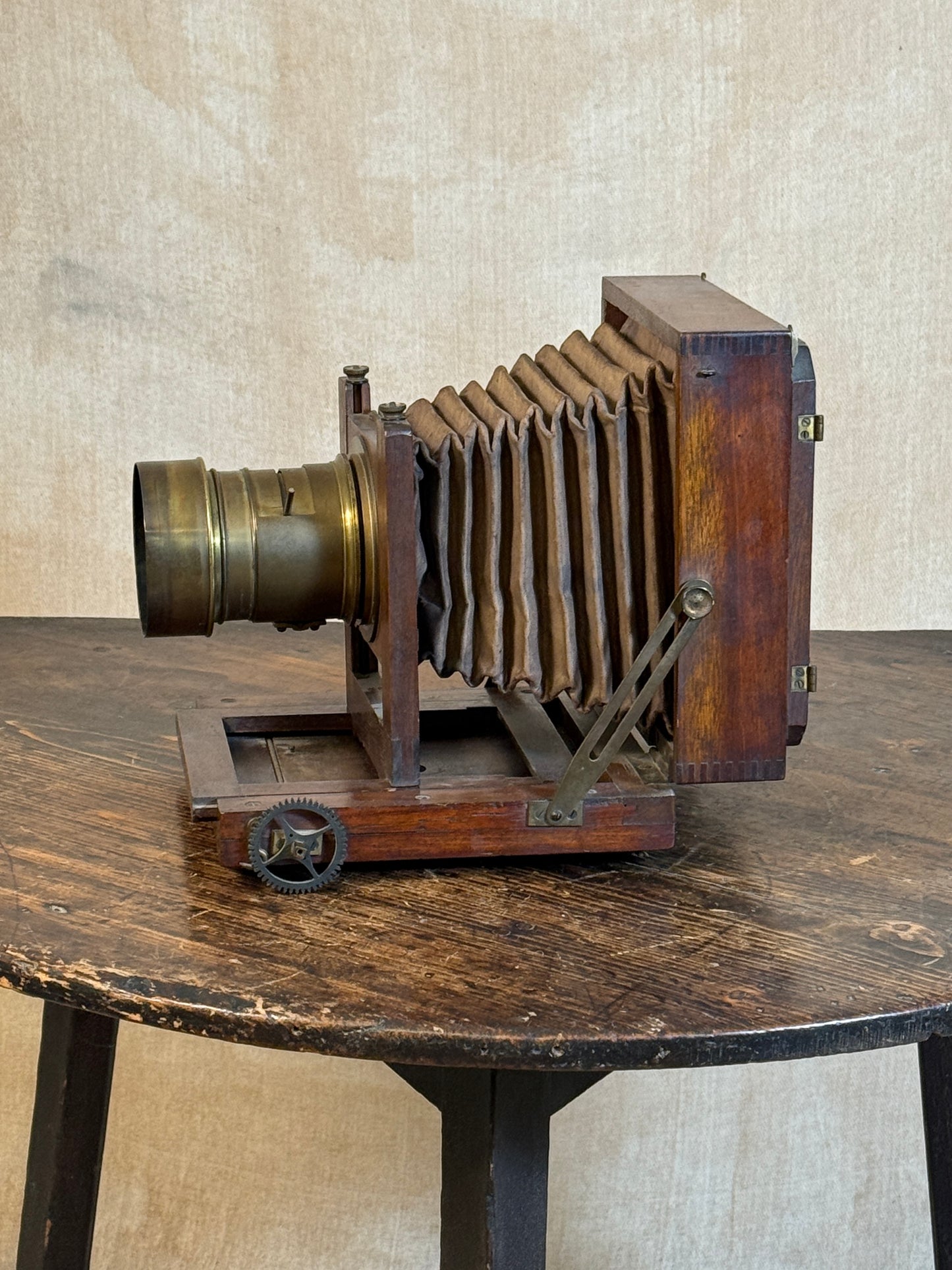 Vintage wooden bellows camera with a brass lens, displayed on a rustic wooden table against a textured beige wall.