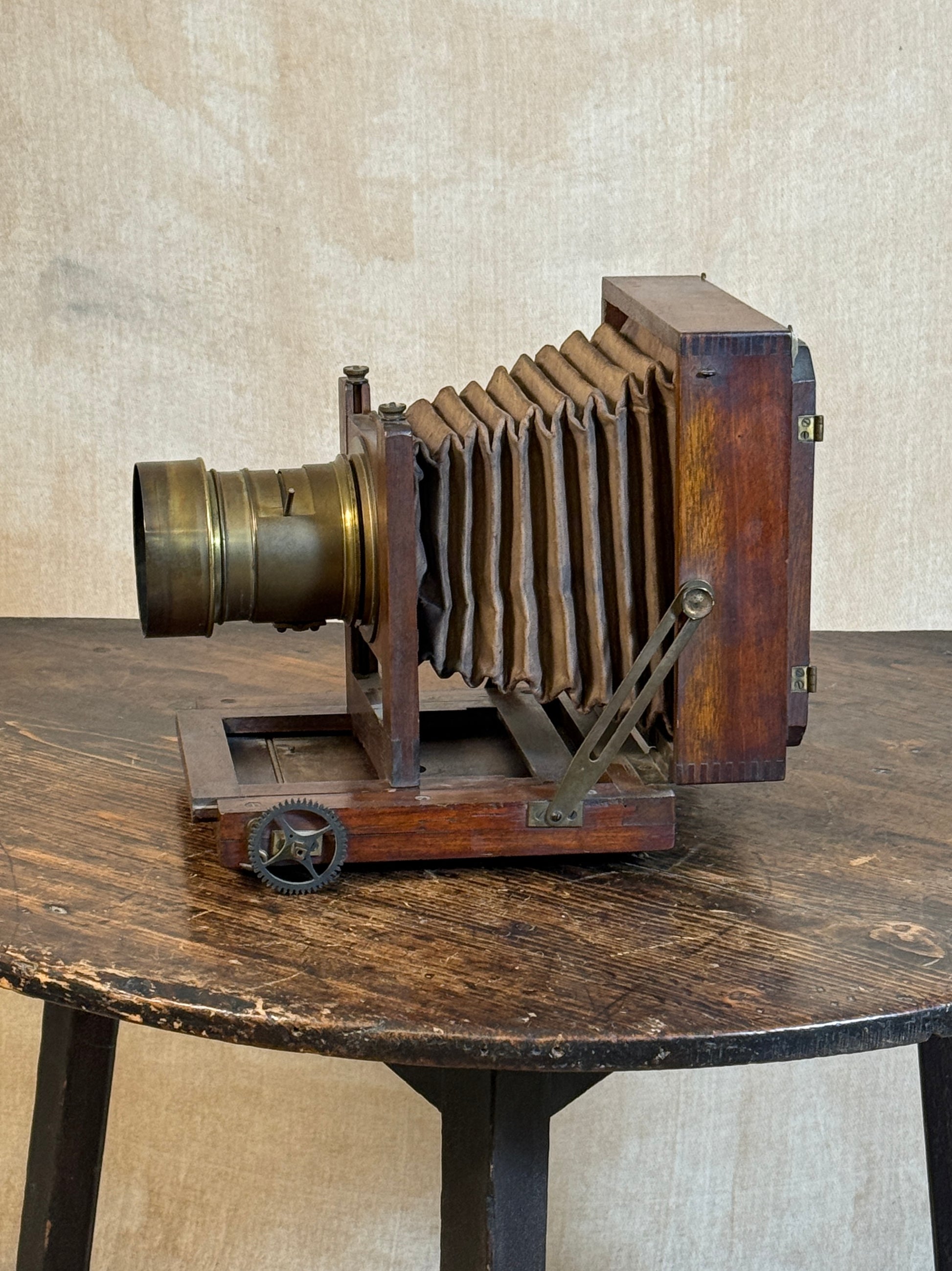 Vintage wooden bellows camera with a brass lens, displayed on a rustic wooden table against a textured beige wall.