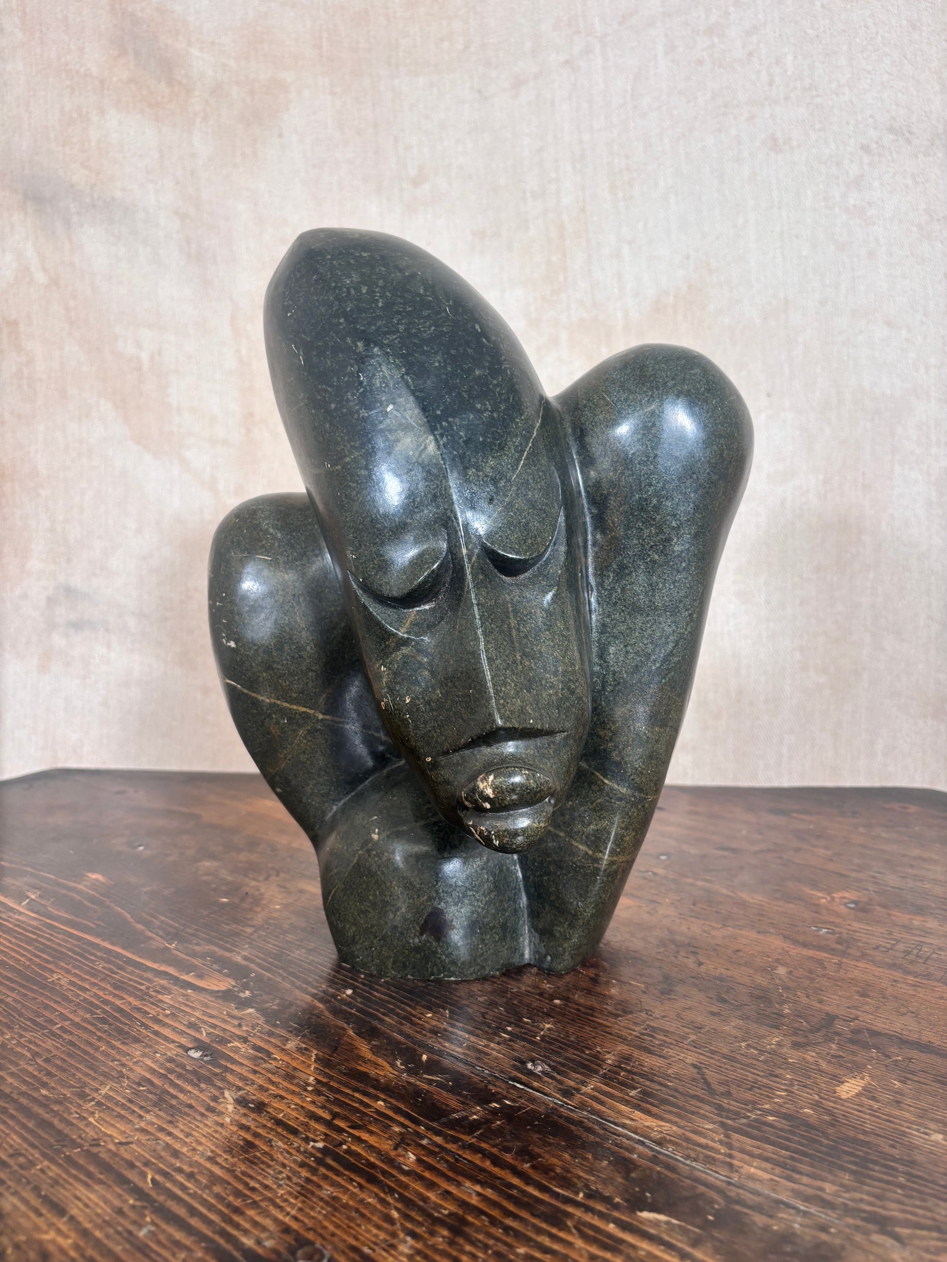 A dark polished stone sculpture of a stylized face resting on its hands, displayed on a wooden surface against a neutral textured background.