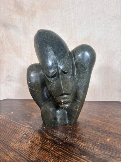 A dark polished stone sculpture of a stylized face resting on its hands, displayed on a wooden surface against a neutral textured background.