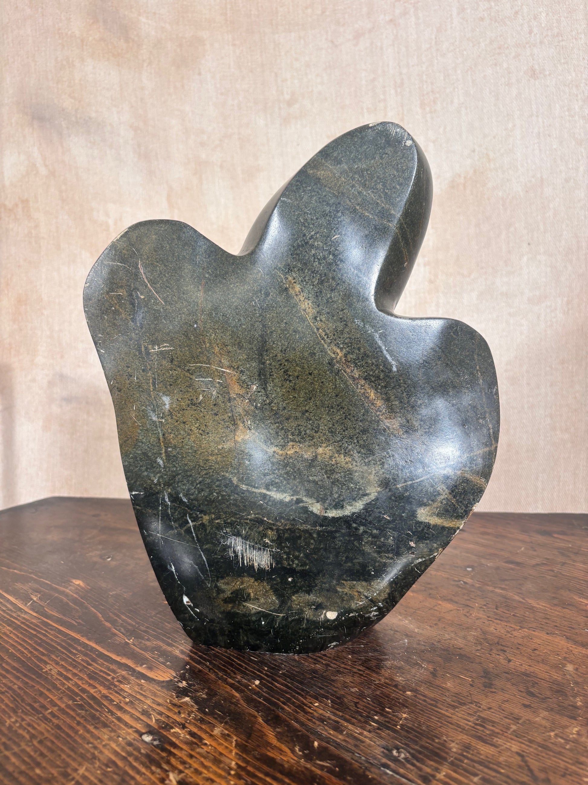 A polished abstract stone sculpture with organic flowing contours, resting on a dark wooden table against a textured beige wall.