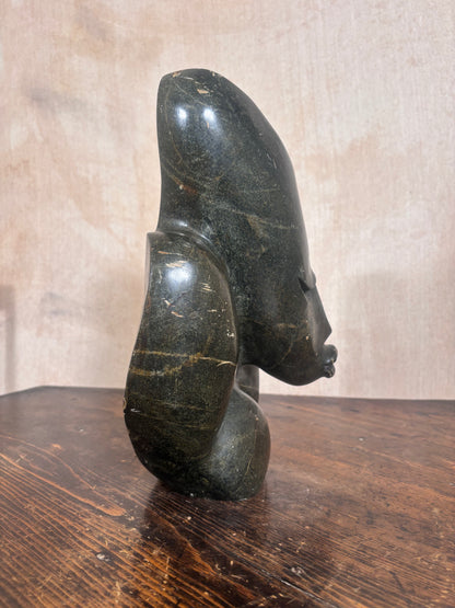 A dark polished stone sculpture of a stylized female head and neck, standing on a wooden surface against a textured beige wall.