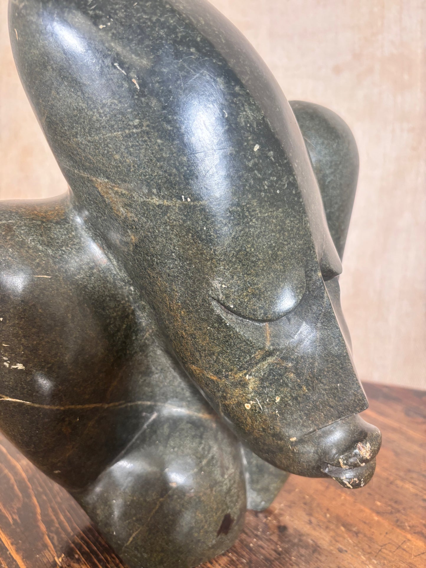 A close-up of a dark polished stone sculpture depicting a stylized animal head, likely a dog or wolf, resting on a wooden surface against a neutral beige background.