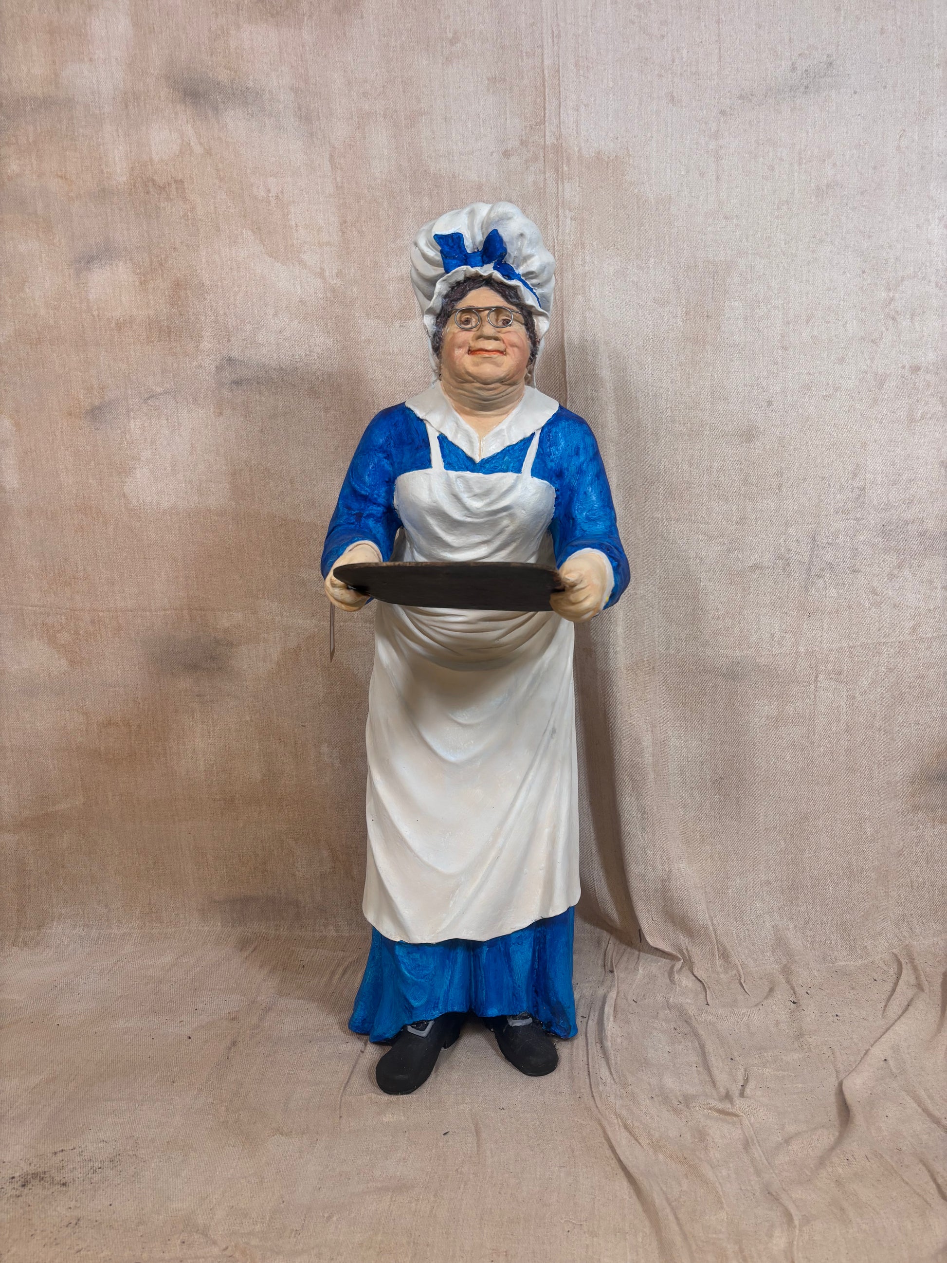 A vintage-style female cook figurine wearing a white apron and blue dress, holding a black serving tray, set against a draped beige fabric backdrop.