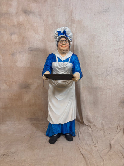 A vintage-style female cook figurine wearing a white apron and blue dress, holding a black serving tray, set against a draped beige fabric backdrop.