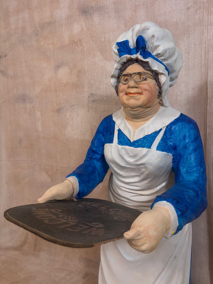 A painted figurine of a woman in a blue and white apron and chef-style hat holding a dark sign, photographed against a textured beige backdrop.