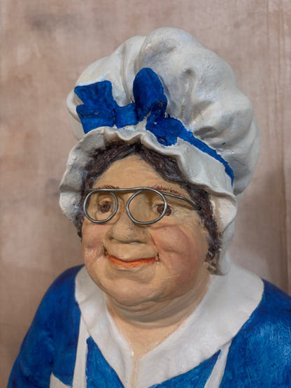 A close-up portrait of a decorative figurine or sculpture depicting an older woman with round glasses, a white ruffled bonnet with a blue bow, and a blue-and-white dress, set against a textured beige background.