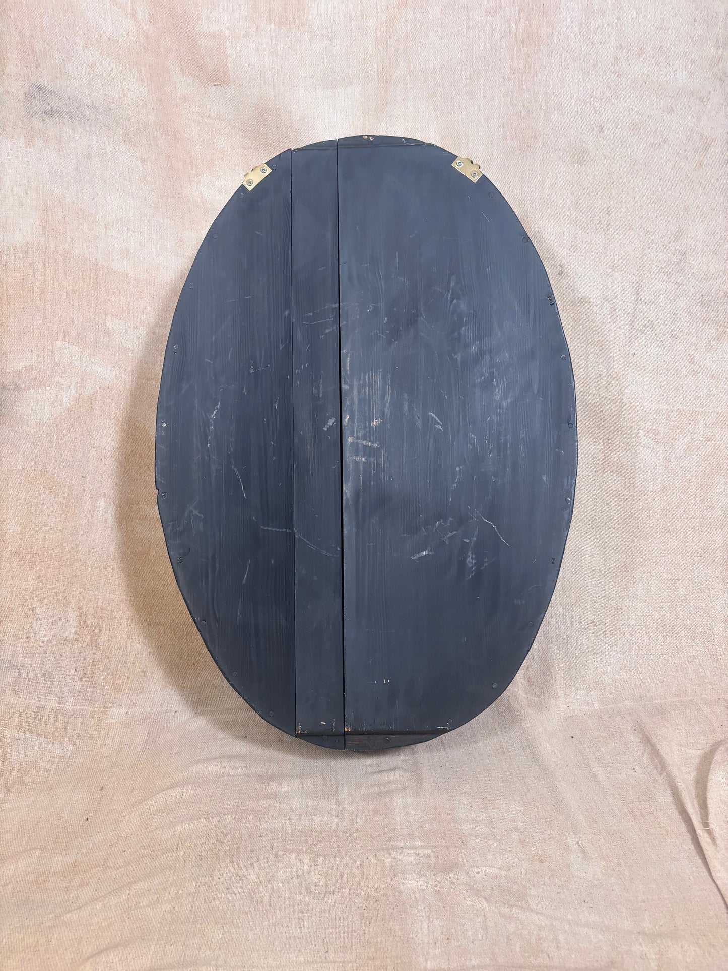 An oval-shaped black wooden box or cabinet with a vertical seam down the center, photographed against a beige fabric backdrop.