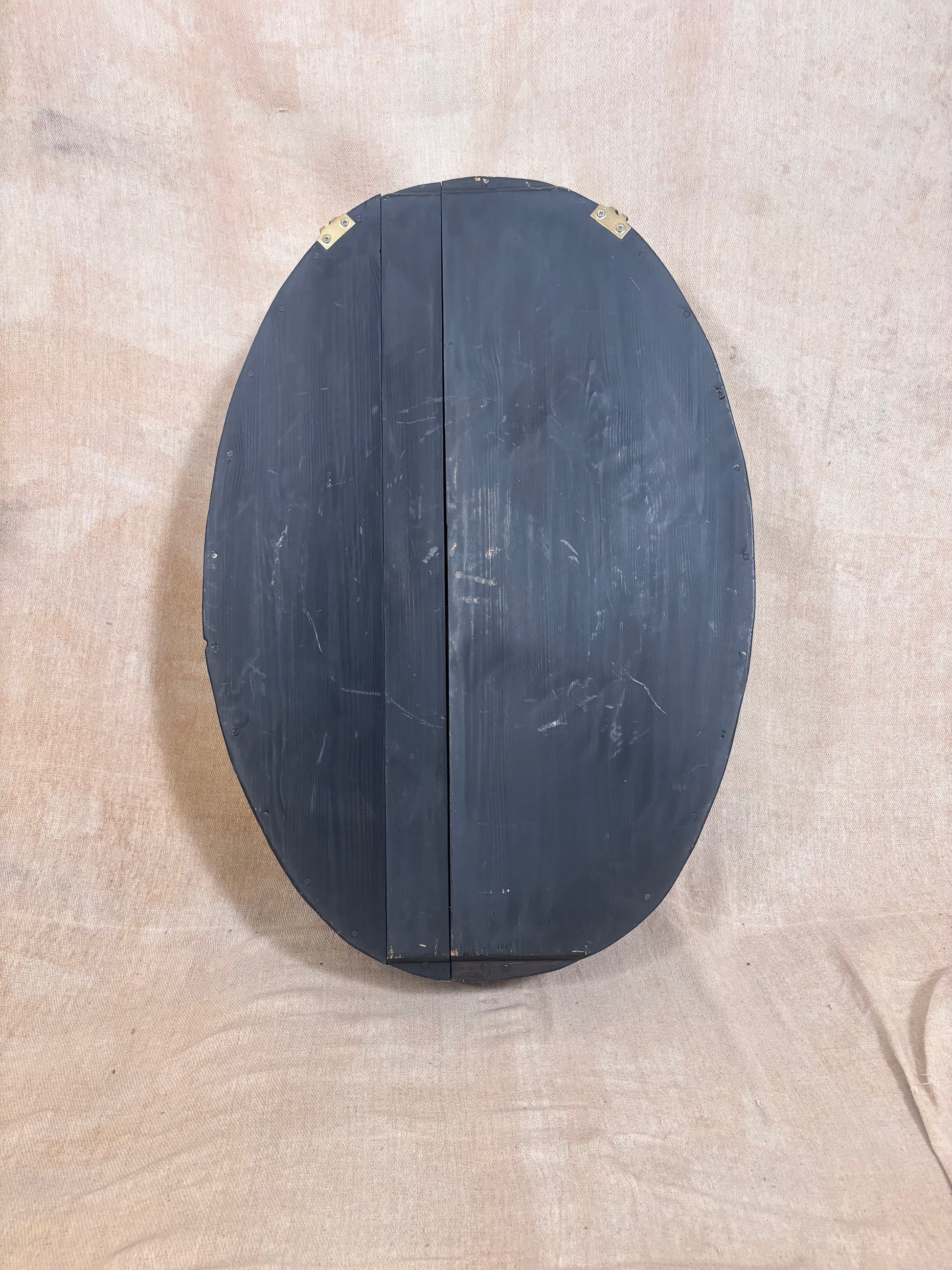 An oval-shaped black wooden box or cabinet with a vertical seam down the center, photographed against a beige fabric backdrop.