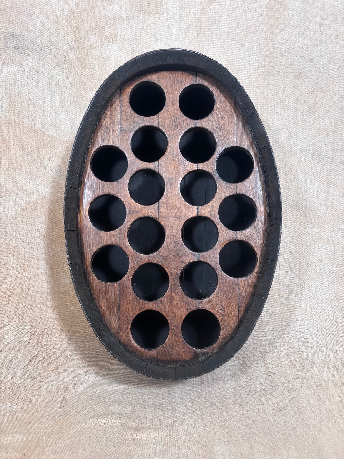 An oval wooden wall-mounted wine bottle holder with a dark outer border and 15 circular cutouts, shown against a light beige textured wall.