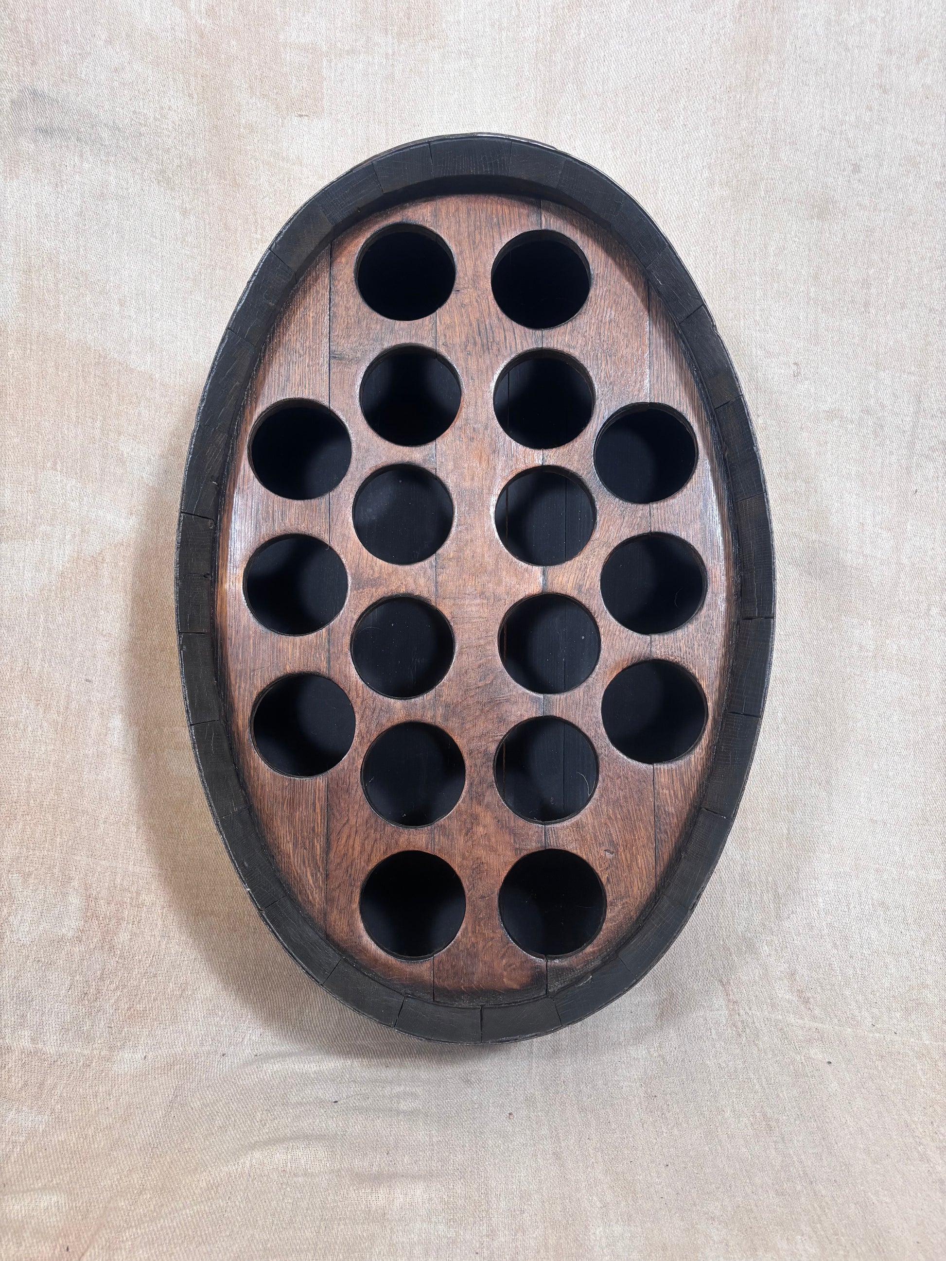An oval wooden wall-mounted wine bottle holder with a dark outer border and 15 circular cutouts, shown against a light beige textured wall.