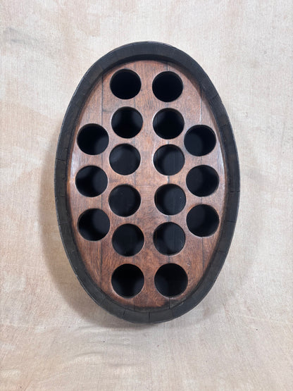 An oval wooden wall-mounted wine bottle holder with a dark outer border and 15 circular cutouts, shown against a light beige textured wall.