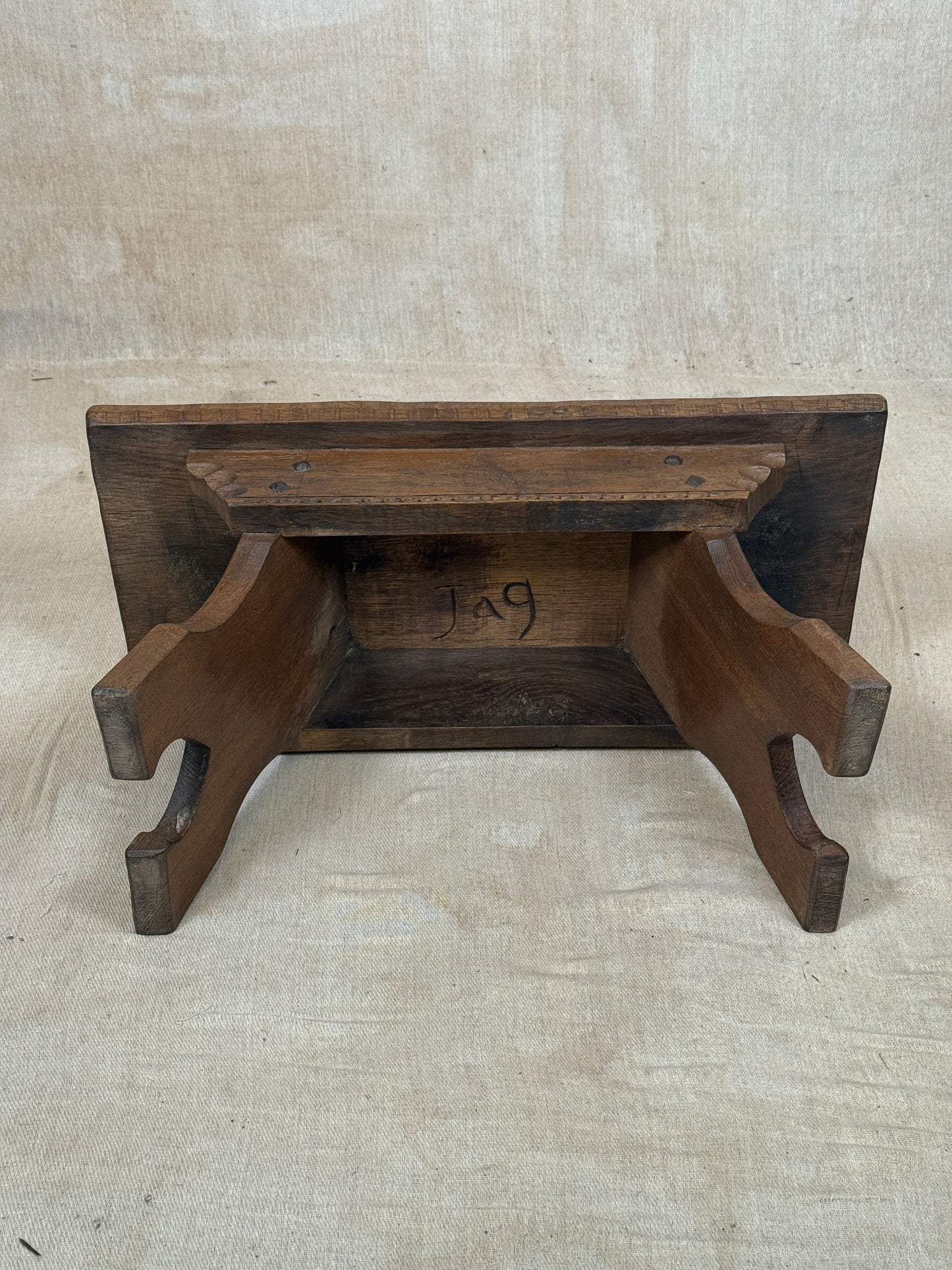 A rustic dark brown wooden shelf or bookcase with a slatted back panel and curved support legs, photographed against a neutral beige fabric backdrop.