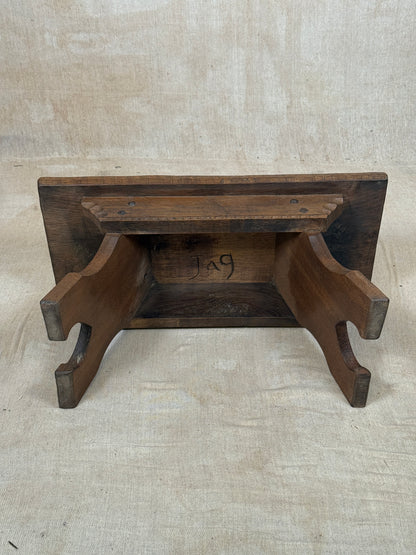 A rustic dark brown wooden shelf or bookcase with a slatted back panel and curved support legs, photographed against a neutral beige fabric backdrop.