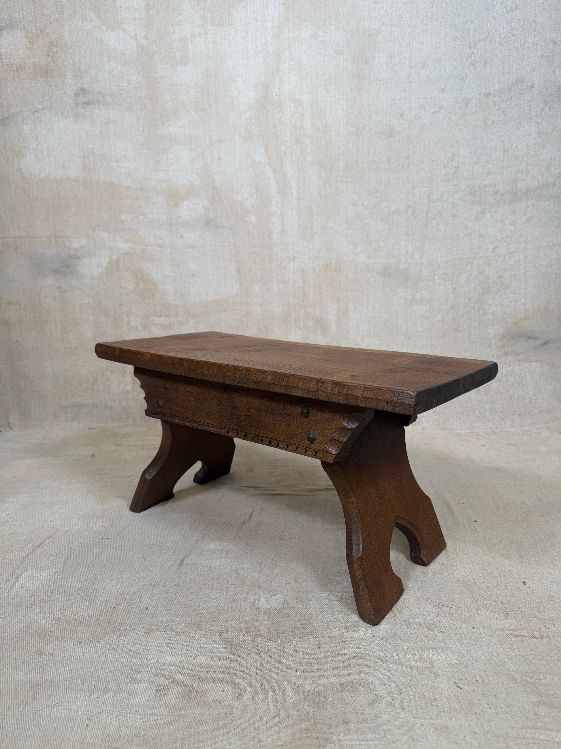 A rustic dark brown wooden bench with a thick rectangular seat and curved, carved legs, set against a textured beige wall and floor.