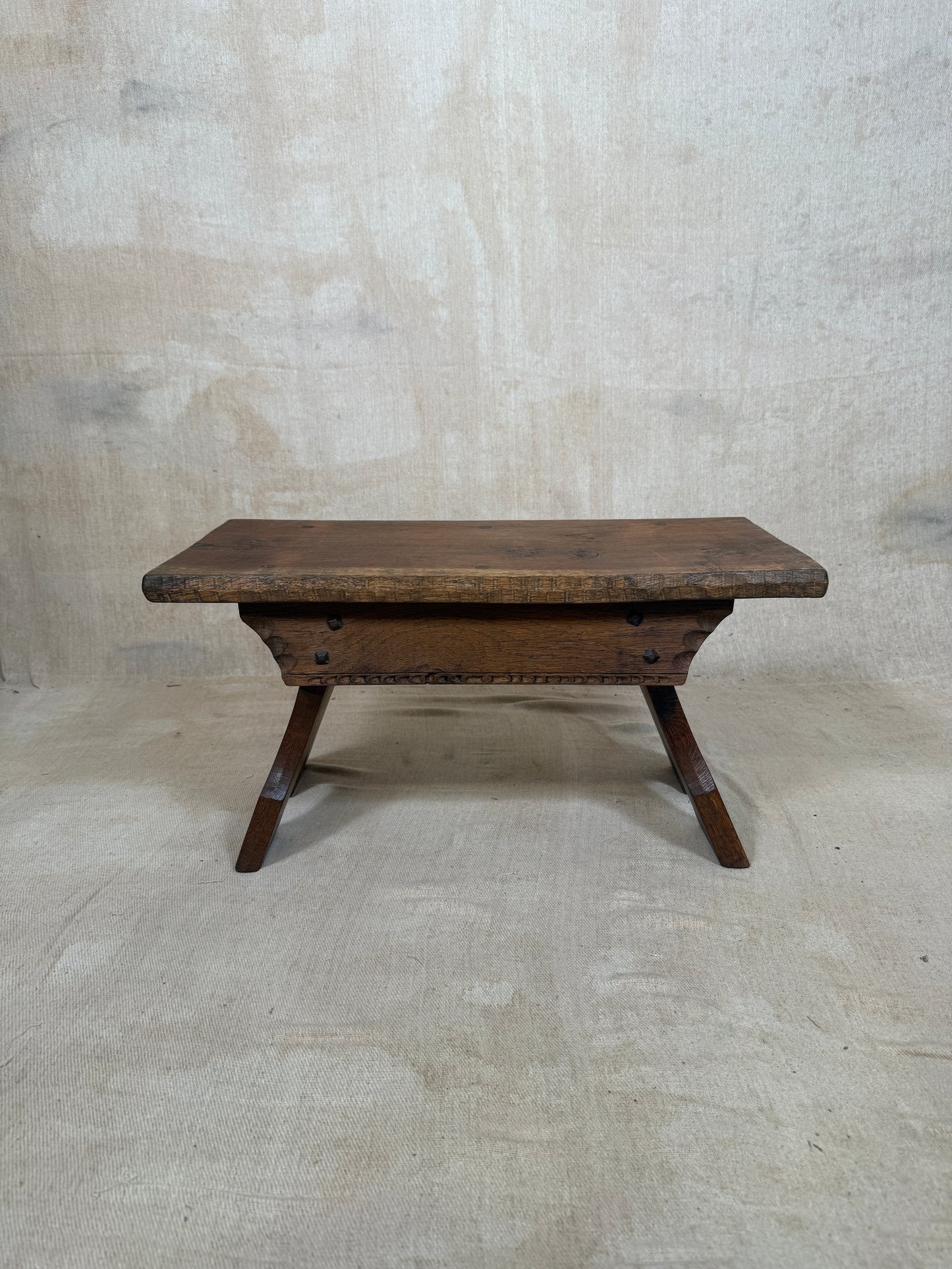A rustic dark brown wooden bench with a thick rectangular seat and angled legs, set against a textured light beige concrete wall and floor.
