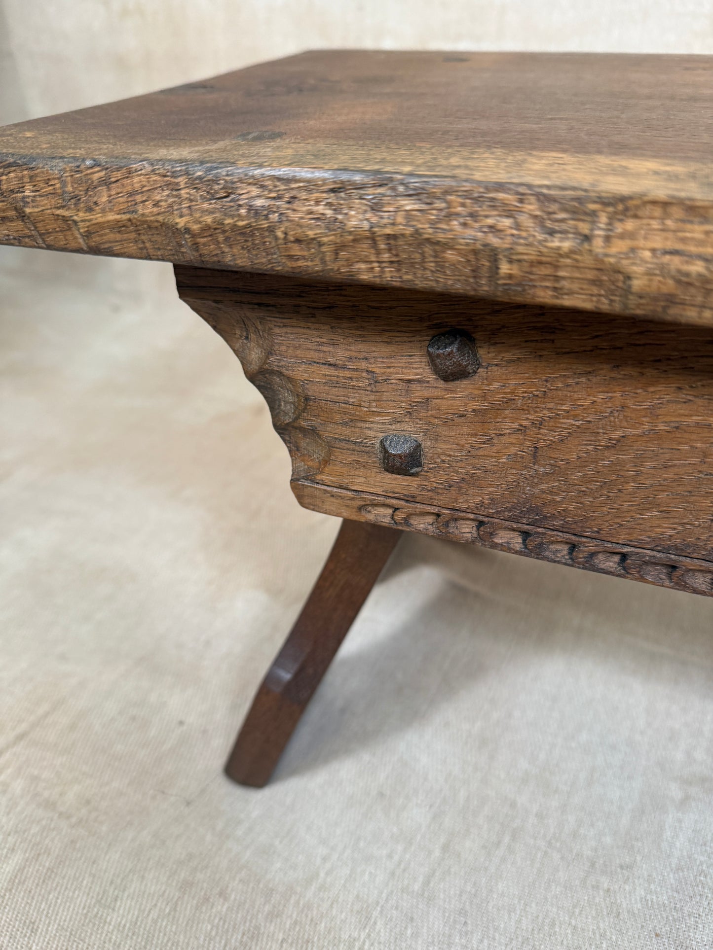 Close-up of an antique wooden table showing the corner joint, carved apron, two dark metal drawer knobs, and a turned leg on a beige fabric background.