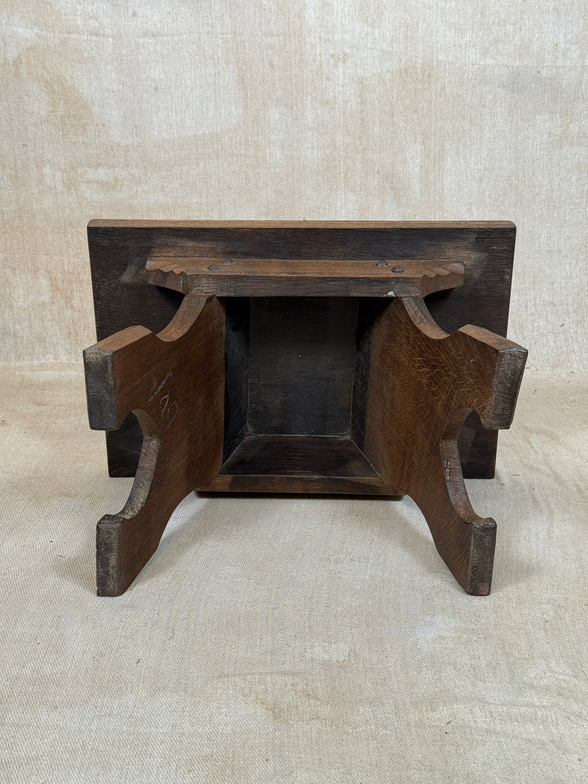 A dark brown wooden stool or bench with curved side supports and a rectangular backrest, photographed against a neutral beige fabric backdrop.