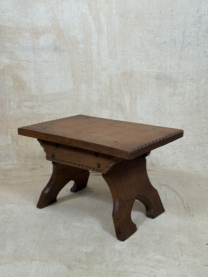 A dark brown wooden table or stool with a rectangular top and four curved legs, photographed against a textured beige fabric backdrop.