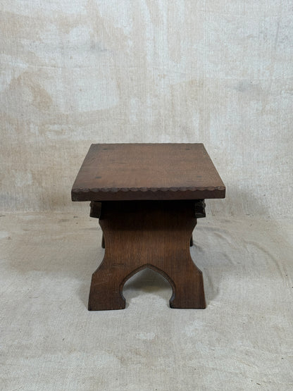 A small dark brown wooden stool with a square top, decorative beaded edge, and a carved arched support base, set against a textured beige fabric backdrop.